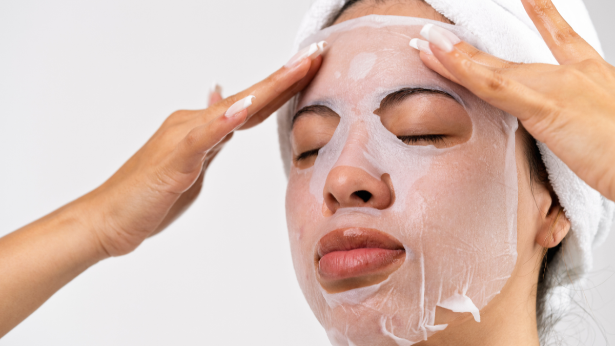 Frau mit weißer Sheet-Maske im Gesicht streicht die Maske an der Stirn glatt.
