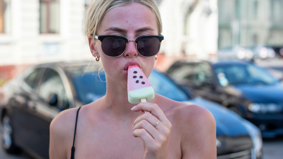 Blonde junge Frau mit Sonnenbrand im Gesicht trägt Sonnenbrille und isst ein Eis
