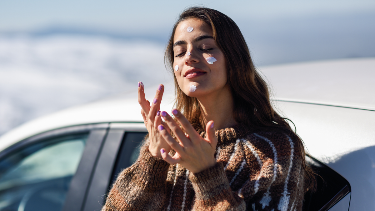 junge Frau steht vor ihrem Auto cremt sich ihr Gesicht ein Schnee im Hintergrund