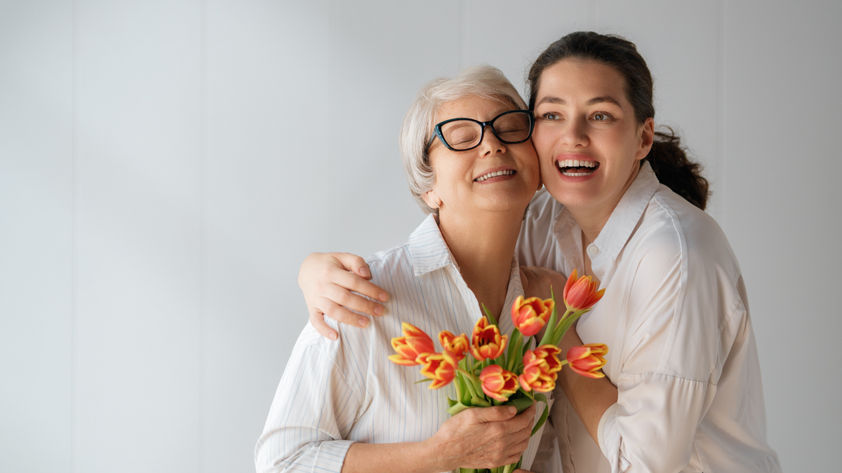 Frau umarmt ihre Mutter, beide lächeln die Mutter hält orangene Tulpen in der Hand