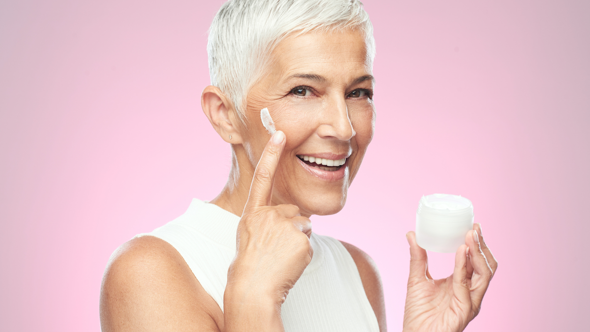 Eine ältere Damen mit kurzen weißen Haaren trägt Feuchtigkeitscreme auf ihrer rechten Wange auf 